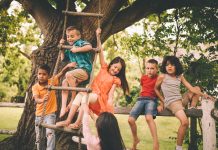 Group of children from mixed racial groups playing in a park on a rustic wooden fence and a rope ladder hanging down from a large tree on a summer day