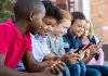 Pupils using mobile phone at the elementary school during recreation time. Group of multiethnic children sitting in a row and typing a message on smartphone. Young boys and girls playing with cellphone.