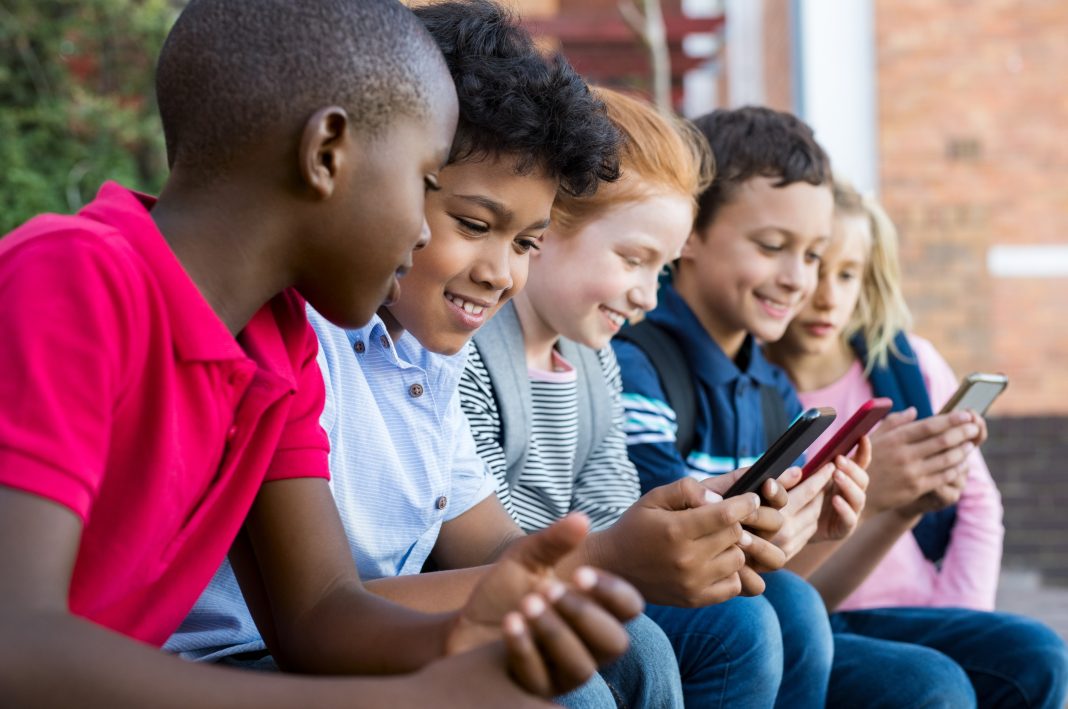 Pupils using mobile phone at the elementary school during recreation time. Group of multiethnic children sitting in a row and typing a message on smartphone. Young boys and girls playing with cellphone.