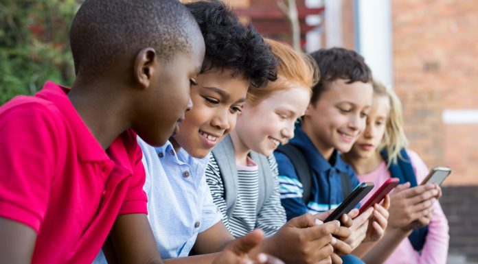 Pupils using mobile phone at the elementary school during recreation time. Group of multiethnic children sitting in a row and typing a message on smartphone. Young boys and girls playing with cellphone.