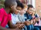 Pupils using mobile phone at the elementary school during recreation time. Group of multiethnic children sitting in a row and typing a message on smartphone. Young boys and girls playing with cellphone.