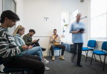 People sitting in a vaccine waiting room, they are waiting to get their COVID-19 vaccine. They are all using their mobile phones. There is a medical professional walking through the room, who is blurred in motion.