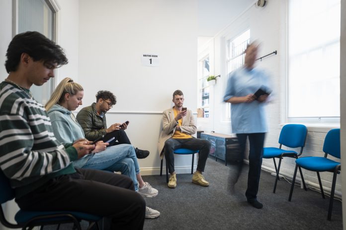 People sitting in a vaccine waiting room, they are waiting to get their COVID-19 vaccine. They are all using their mobile phones. There is a medical professional walking through the room, who is blurred in motion.