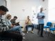 People sitting in a vaccine waiting room, they are waiting to get their COVID-19 vaccine. They are all using their mobile phones. There is a medical professional walking through the room, who is blurred in motion.