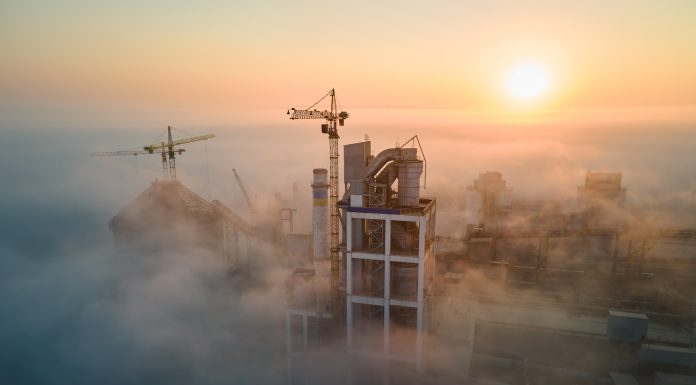 Aerial view of cement factory with high concrete plant structure and tower crane at industrial production site on foggy morning. Manufacture and global industry concept.