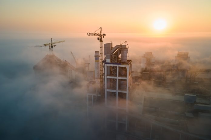 Aerial view of cement factory with high concrete plant structure and tower crane at industrial production site on foggy morning. Manufacture and global industry concept.