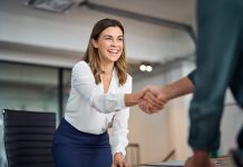 Happy mid aged business woman manager handshaking greeting client in office. Smiling female executive making successful deal with partner shaking hand at work standing at meeting table.