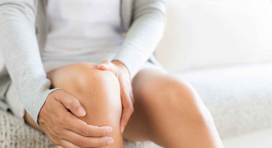 Closeup young woman sitting on sofa and feeling knee pain and she massage her knee at home. Healthcare and medical concept.