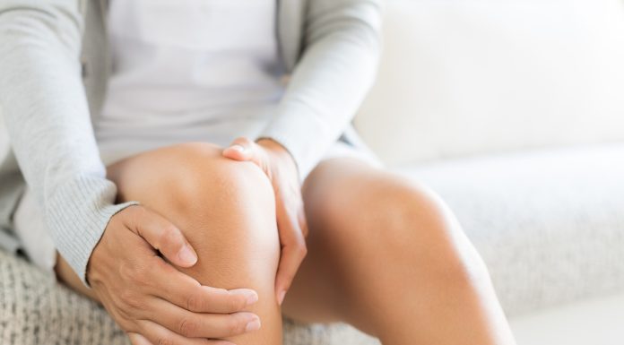 Closeup young woman sitting on sofa and feeling knee pain and she massage her knee at home. Healthcare and medical concept.