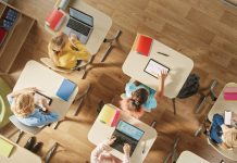 Top View Shot in Elementary School Computer Science Classroom: Children Sitting at their School Desk Using Personal Computers and Digital Tablets for Assignments.