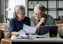 old retired asian senior couple checking and calculate financial billing together on sofa involved in financial paperwork, paying taxes online using e-banking laptop at living room home background