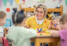 A small group of preschool children sit with their teacher at a table as they learn through play. They are each dressed casually and are focused on the activity.