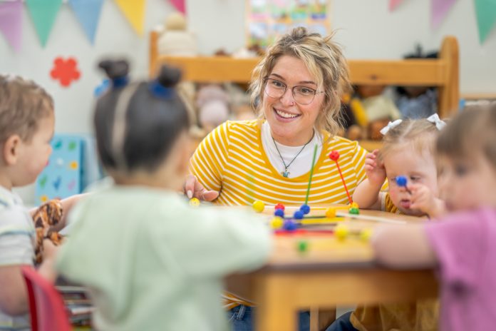 A small group of preschool children sit with their teacher at a table as they learn through play. They are each dressed casually and are focused on the activity.