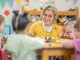 A small group of preschool children sit with their teacher at a table as they learn through play. They are each dressed casually and are focused on the activity.