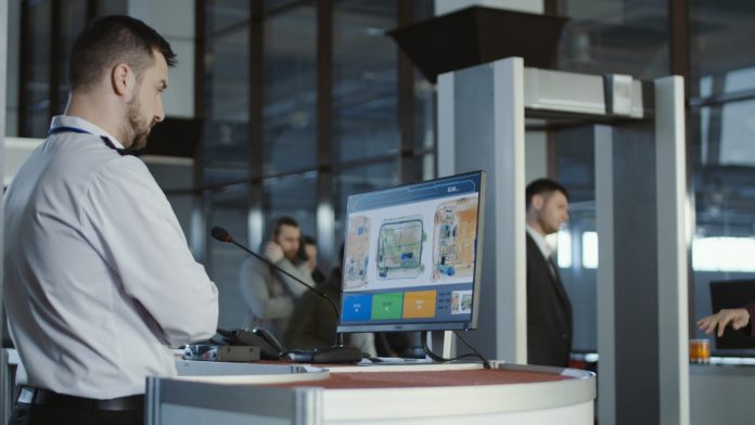 Man in uniform standing at counter at checking point and watching at monitor with x-ray of luggage.