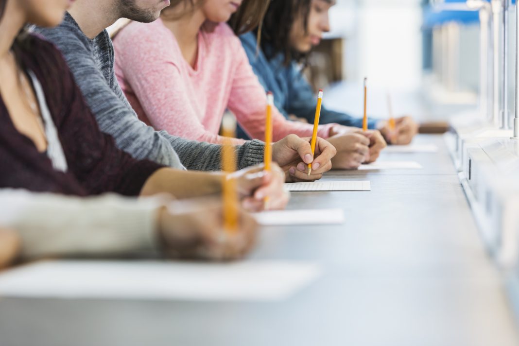 Cropped view of a multiracial group of young men and women sitting in a row at a table, writing with pencils on paper. They are taking a test or filling out an application. Focus is on the hand of the young man in the middle in the gray shirt.