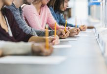 Cropped view of a multiracial group of young men and women sitting in a row at a table, writing with pencils on paper. They are taking a test or filling out an application. Focus is on the hand of the young man in the middle in the gray shirt.