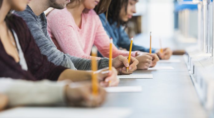 Cropped view of a multiracial group of young men and women sitting in a row at a table, writing with pencils on paper. They are taking a test or filling out an application. Focus is on the hand of the young man in the middle in the gray shirt.