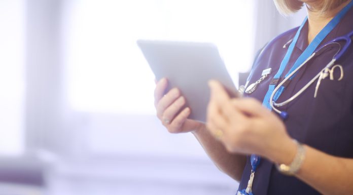 Female surgeon typing on digital tablet in hospital or surgery. She is wearing a dark blue nurse’s top and has her stethoscope around her neck along with her nhs lanyard. She is looking at her patients records on her digital tablet and sending emails.