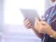 Female surgeon typing on digital tablet in hospital or surgery. She is wearing a dark blue nurse’s top and has her stethoscope around her neck along with her nhs lanyard. She is looking at her patients records on her digital tablet and sending emails.