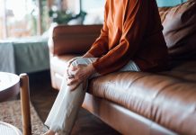 Cropped shot of a distraught senior Asian woman feeling unwell, suffering from pain in leg while sitting on sofa in the living room at home. Elderly and health issues concept