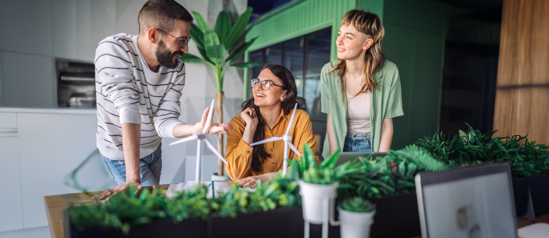 Three young adults, two females and one male, actively brainstorm and discuss wind turbine designs together at a desk filled with plants in a modern, collaborative office space.