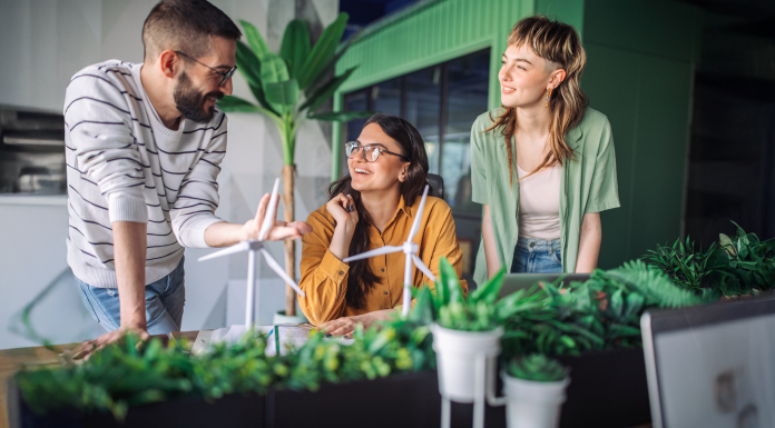 Three young adults, two females and one male, actively brainstorm and discuss wind turbine designs together at a desk filled with plants in a modern, collaborative office space.