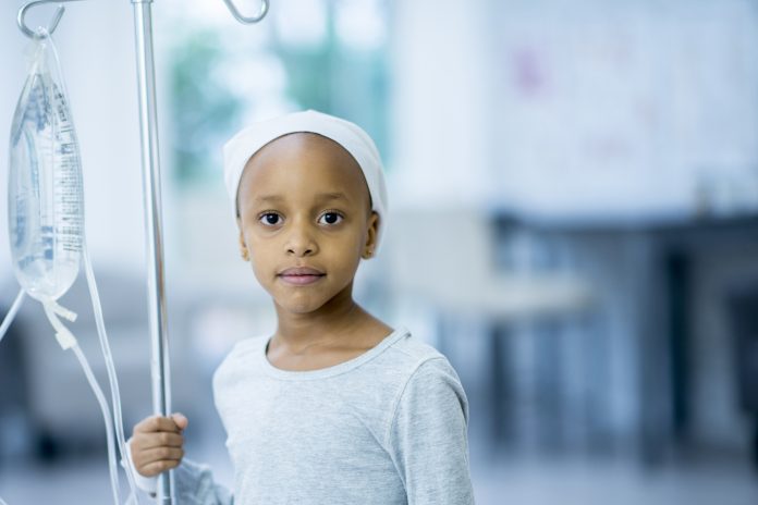 A young girl with cancer is indoors in a hospital. She is holding her IV and staring at the camera.