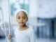 A young girl with cancer is indoors in a hospital. She is holding her IV and staring at the camera.