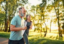 Cardio training outdoors. Side view of caucasian grey bearded man and beautiful old woman running along summer park with sunlight on background. Cheerful retired people leading active lifestyle.