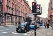 A cab and a bus, stopped at a traffic light on a Manchester street. Perspective view of large brick wall in background.