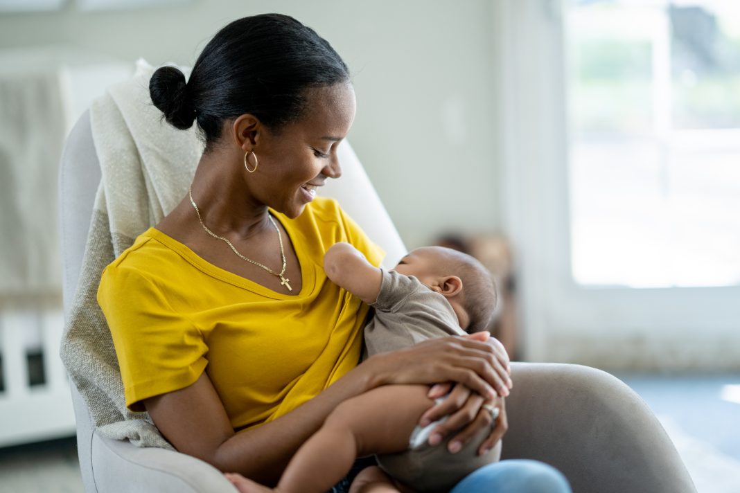 A new Mother sits in her baby's nursery with the infant in her arms and held to her chest, as she nurses him. She is dressed casually in a yellow t-shirt and looking down at the baby with a smile.