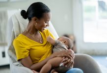 A new Mother sits in her baby's nursery with the infant in her arms and held to her chest, as she nurses him. She is dressed casually in a yellow t-shirt and looking down at the baby with a smile.