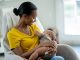 A new Mother sits in her baby's nursery with the infant in her arms and held to her chest, as she nurses him. She is dressed casually in a yellow t-shirt and looking down at the baby with a smile.