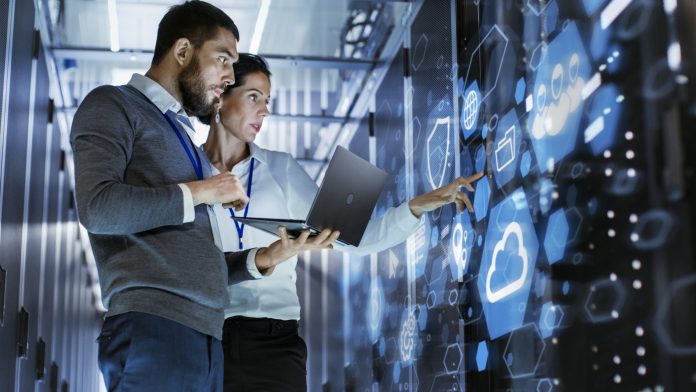 Male IT Specialist Holds Laptop and Discusses Work with Female Server Technician. They're Standing in Data Center, Rack Server Cabinet with Cloud Server Icon and Visualization.