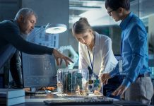 Team of Computer Engineers Lean on the Desk and Choose Printed Circuit Boards to Work with, Computer Shows Programming in Progress. In The Background Technologically Advanced Scientific Research Center.