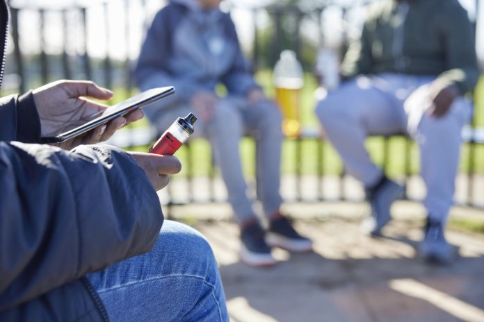 Close Up Of Teenagers With Mobile Phone Vaping and Drinking Alcohol In Park