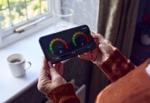 Senior Woman With Smart Meter Trying To Keep Warm By Radiator During Cost Of Living Energy Crisis