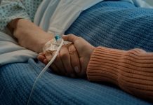 Hospital, holding hands and bed with love, support and care together in a clinic after surgery. Senior, healthcare and cancer patient with empathy, kindness and caregiver with compassion with people