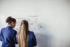 Two teenage school girls standing in front of a large whiteboard side by side solving a mathematics equation on the board. Back view