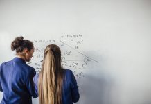 Two teenage school girls standing in front of a large whiteboard side by side solving a mathematics equation on the board. Back view