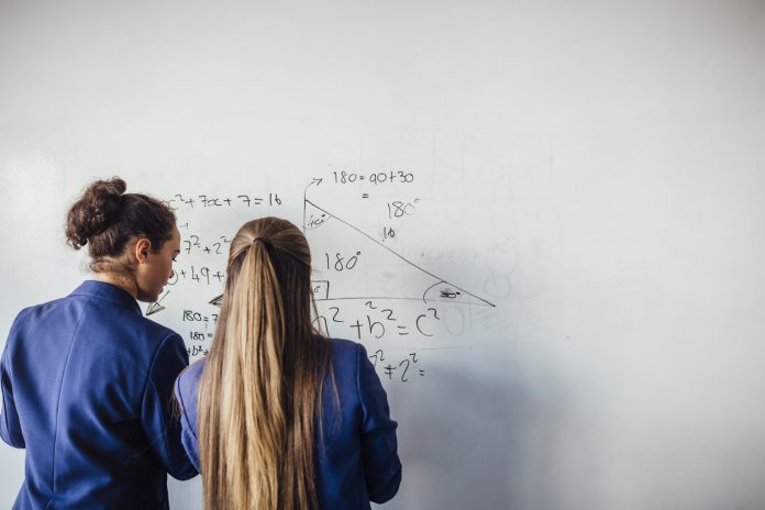 Two teenage school girls standing in front of a large whiteboard side by side solving a mathematics equation on the board. Back view