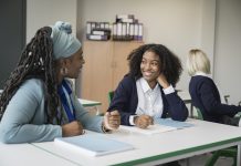 Cheerful instructor in early 50s sitting at desk next to multiracial student, talking and smiling as they discuss her writing assignment in secondary classroom.