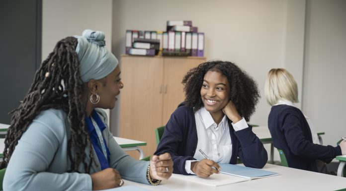 Cheerful instructor in early 50s sitting at desk next to multiracial student, talking and smiling as they discuss her writing assignment in secondary classroom.