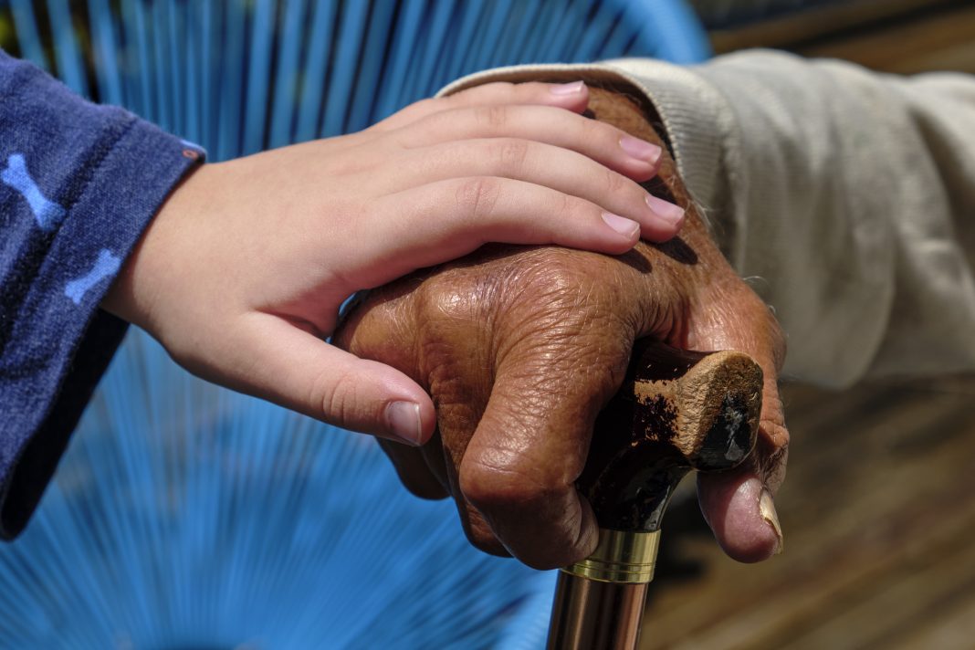 Grandchild holding grandparent's hand. Grandfather leaning on a walking stick. Dark skin toned grandfather with light skin toned grandchild. Mixed race family.