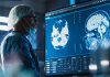 Medical Hospital Research Laboratory: Caucasian Male Neurosurgeon Looking At TV Screen With Brain MRI Scans Of Patient, Analyzing, Preparing For Complicated Neurosurgery, Wearing Surgical Gown.