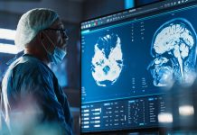 Medical Hospital Research Laboratory: Caucasian Male Neurosurgeon Looking At TV Screen With Brain MRI Scans Of Patient, Analyzing, Preparing For Complicated Neurosurgery, Wearing Surgical Gown.