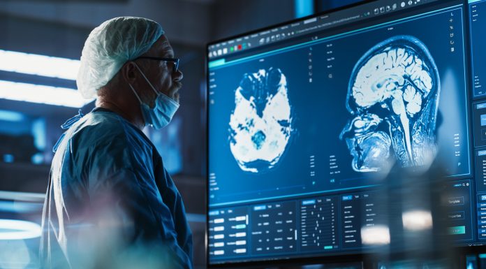 Medical Hospital Research Laboratory: Caucasian Male Neurosurgeon Looking At TV Screen With Brain MRI Scans Of Patient, Analyzing, Preparing For Complicated Neurosurgery, Wearing Surgical Gown.